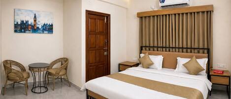 Une chambre d’hôtel comprenant un lit, une tête de lit en bois, une table de chevet, une table avec des chaises, un tableau représentant un paysage urbain et un appareil de climatisation.