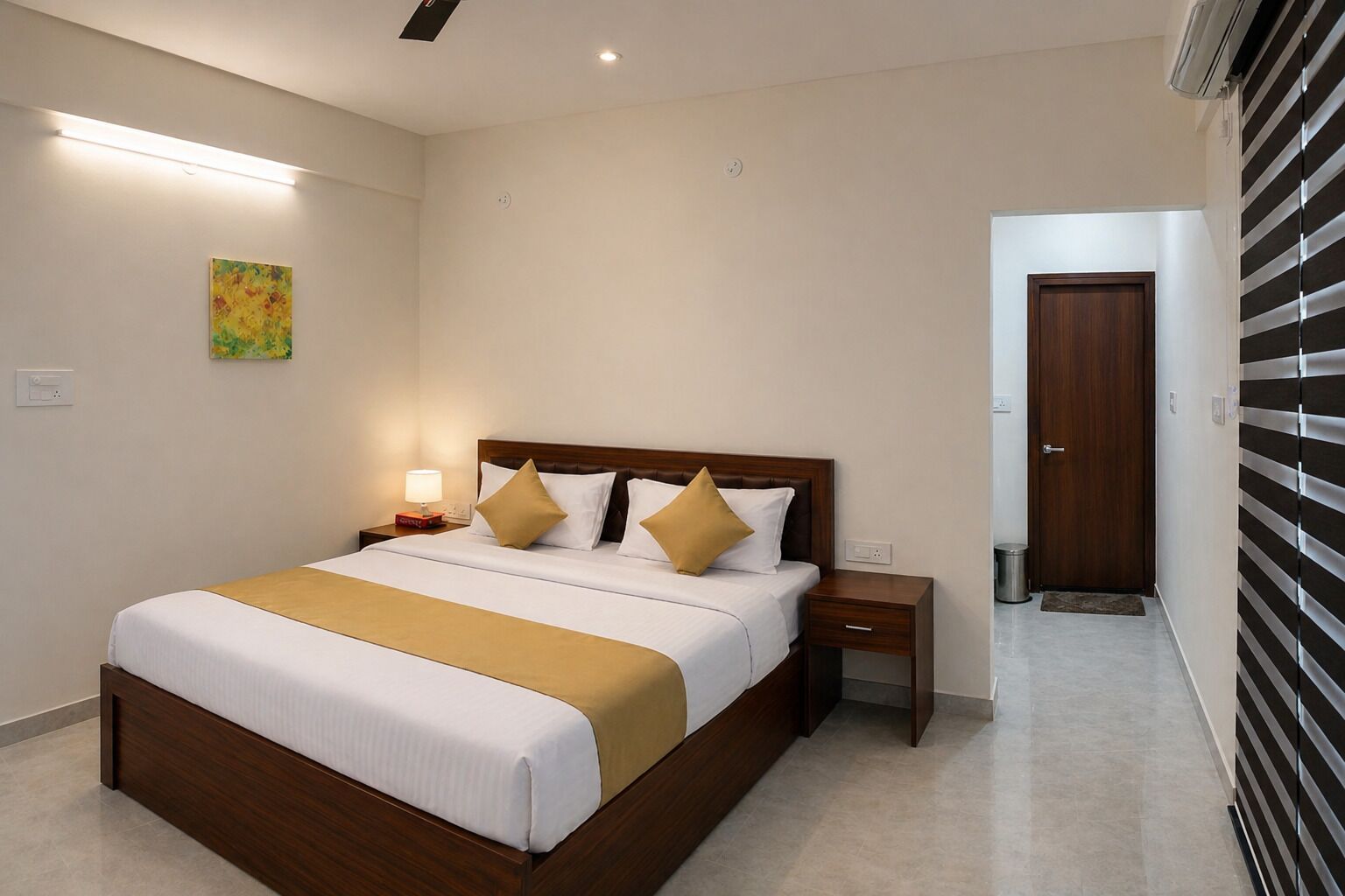 Une chambre d’hôtel avec un grand lit, des tables de chevet, une petite table, une lampe fixée au mur, une porte et une fenêtre avec des stores.