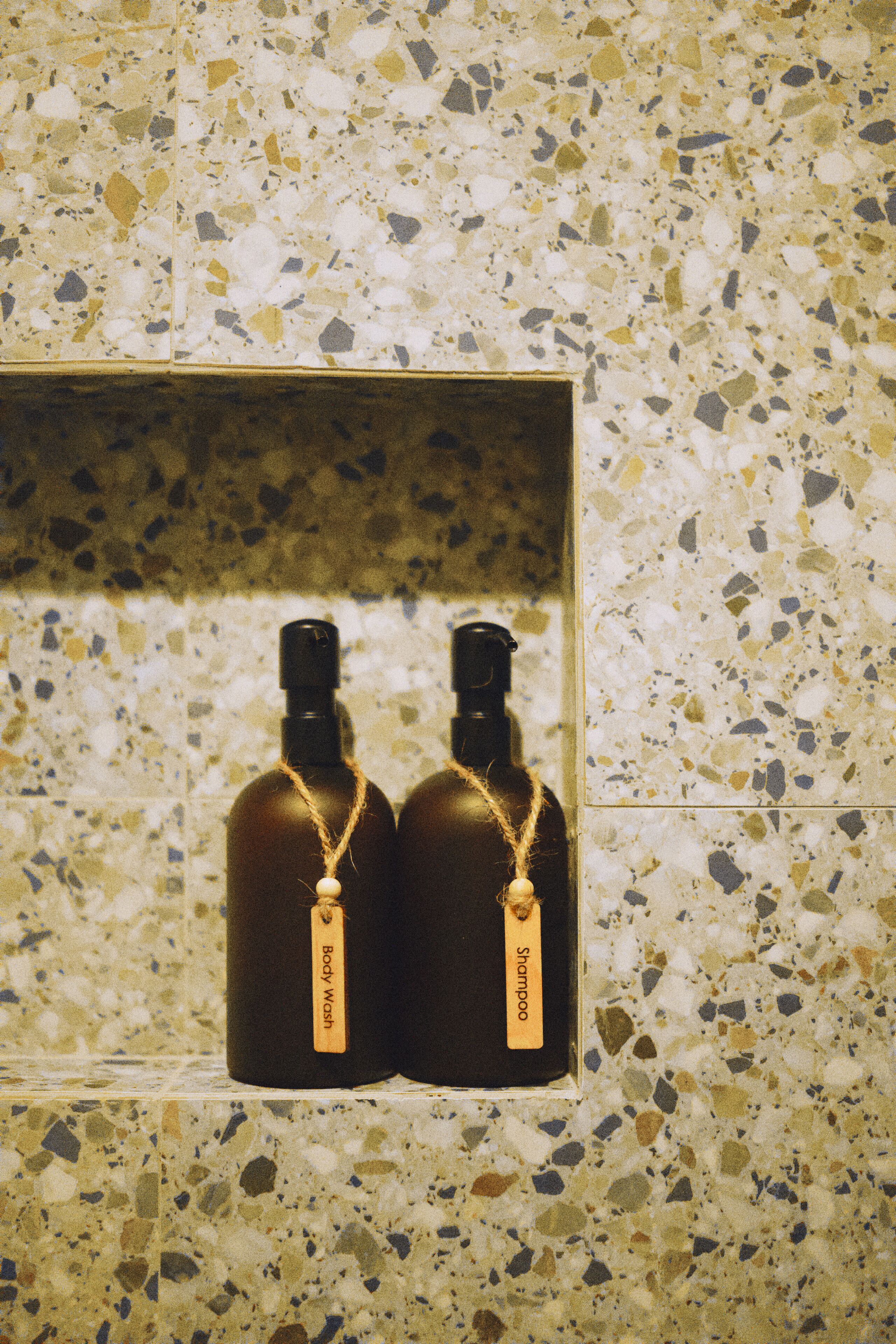 Deux bouteilles de gel douche dans une boîte transparente, le tout placé devant un mur carrelé à motifs.