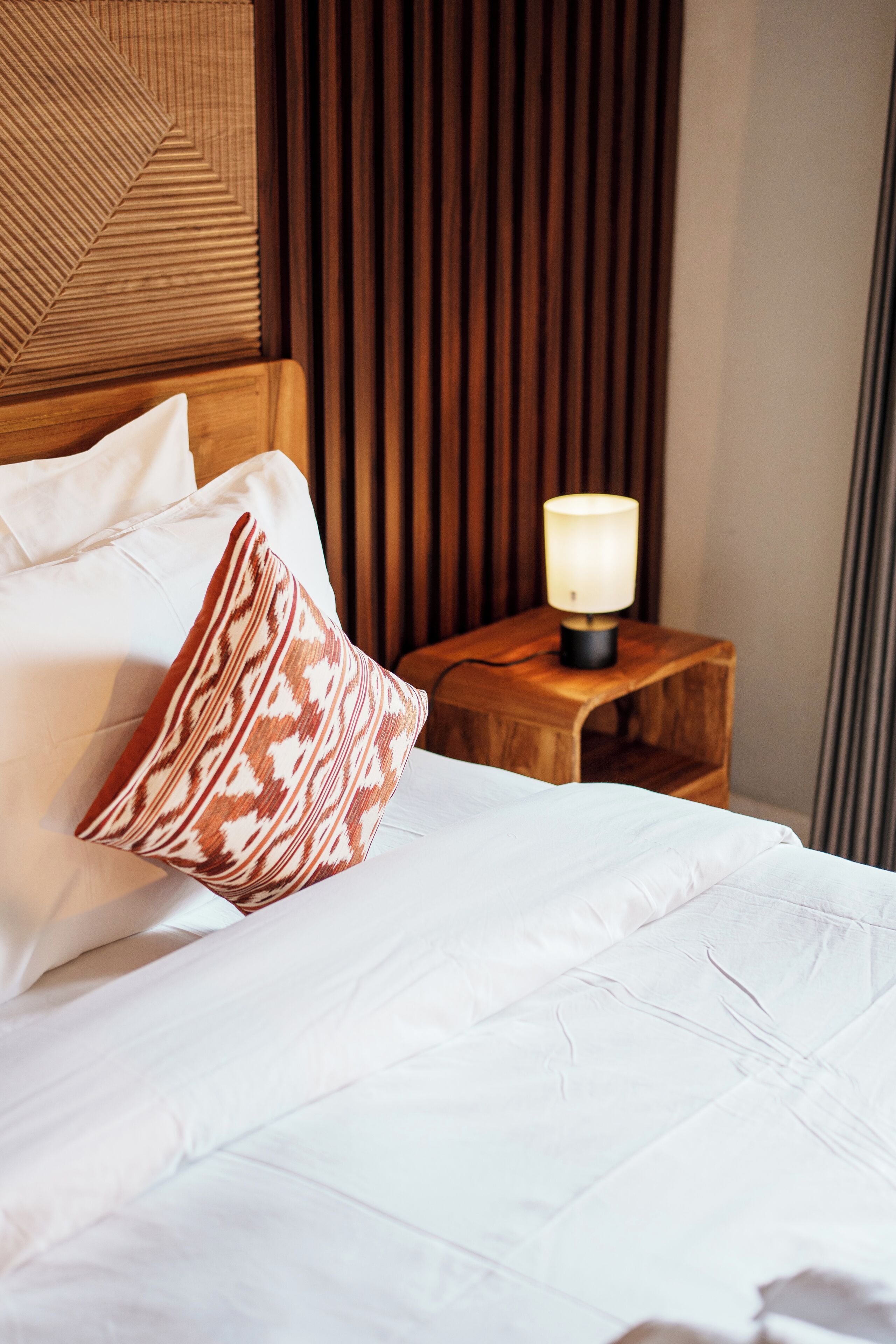 A neatly made bed with white linens and a patterned pillow, a wooden nightstand with a lamp, and a wooden headboard with a geometric design.