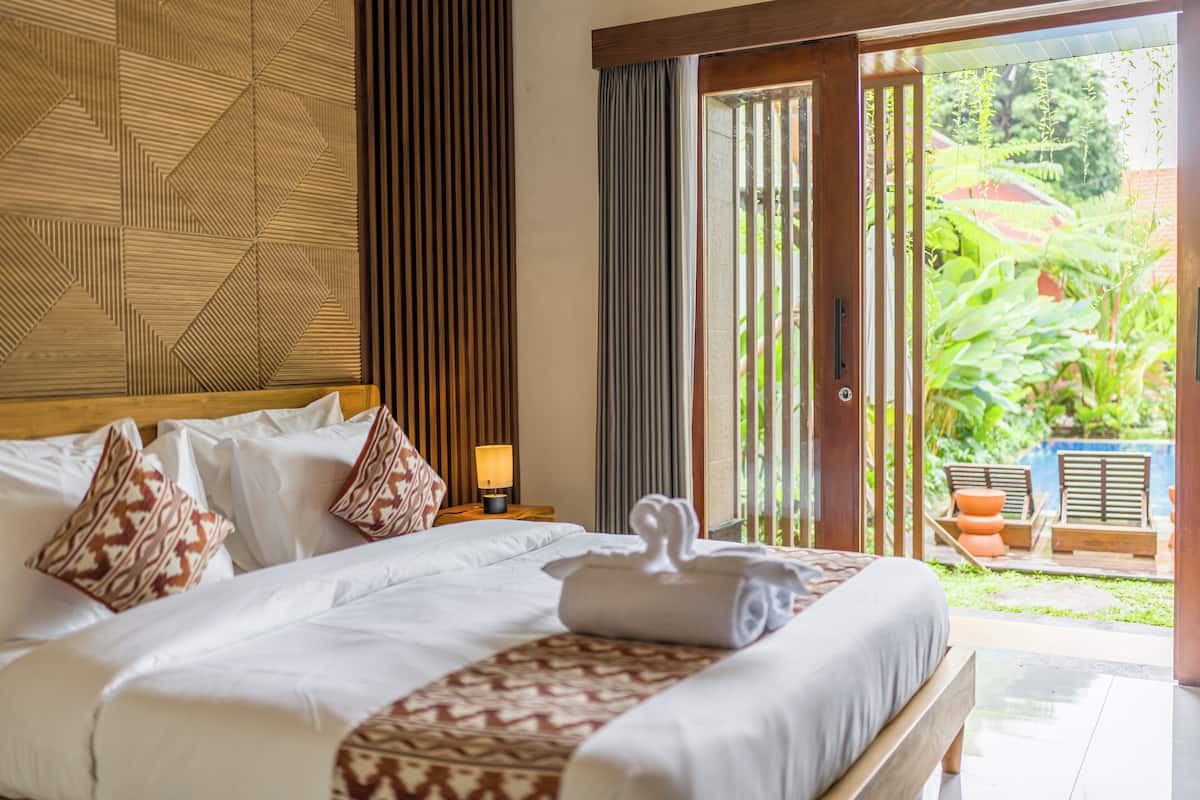 A hotel room with a large bed, a wooden headboard, a nightstand with a lamp, and a view of a garden through a sliding door.
