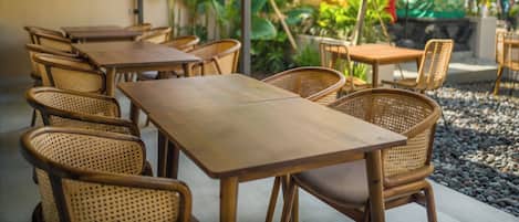 A row of wooden tables and wicker chairs in an outdoor cafe setting with plants and umbrellas in the background.