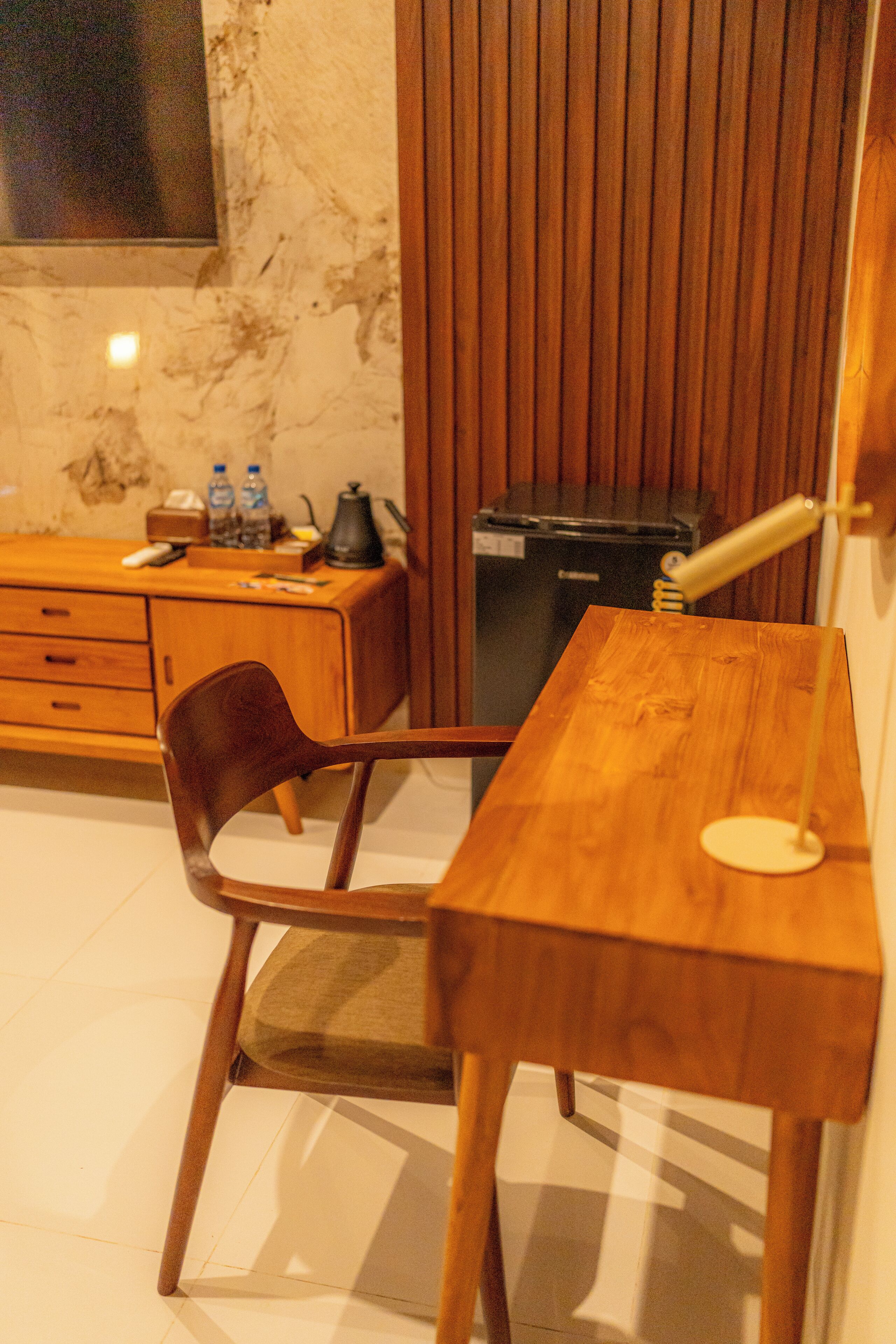 A wooden desk with a chair, a lamp, and a trash bin in a room with a wooden cabinet and a television on the wall.
