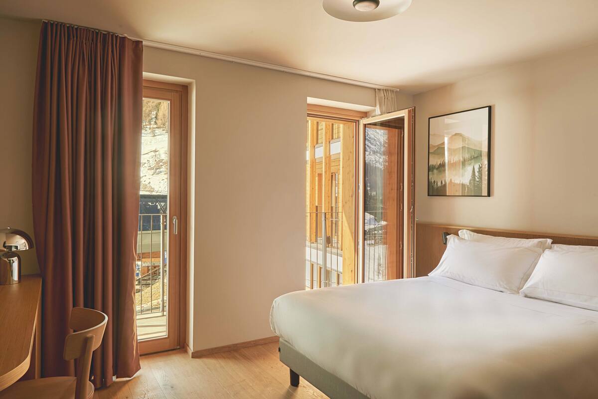 Un dormitorio con cama, escritorio y silla. Hay dos ventanas con cortinas, y un cuadro en la pared.
