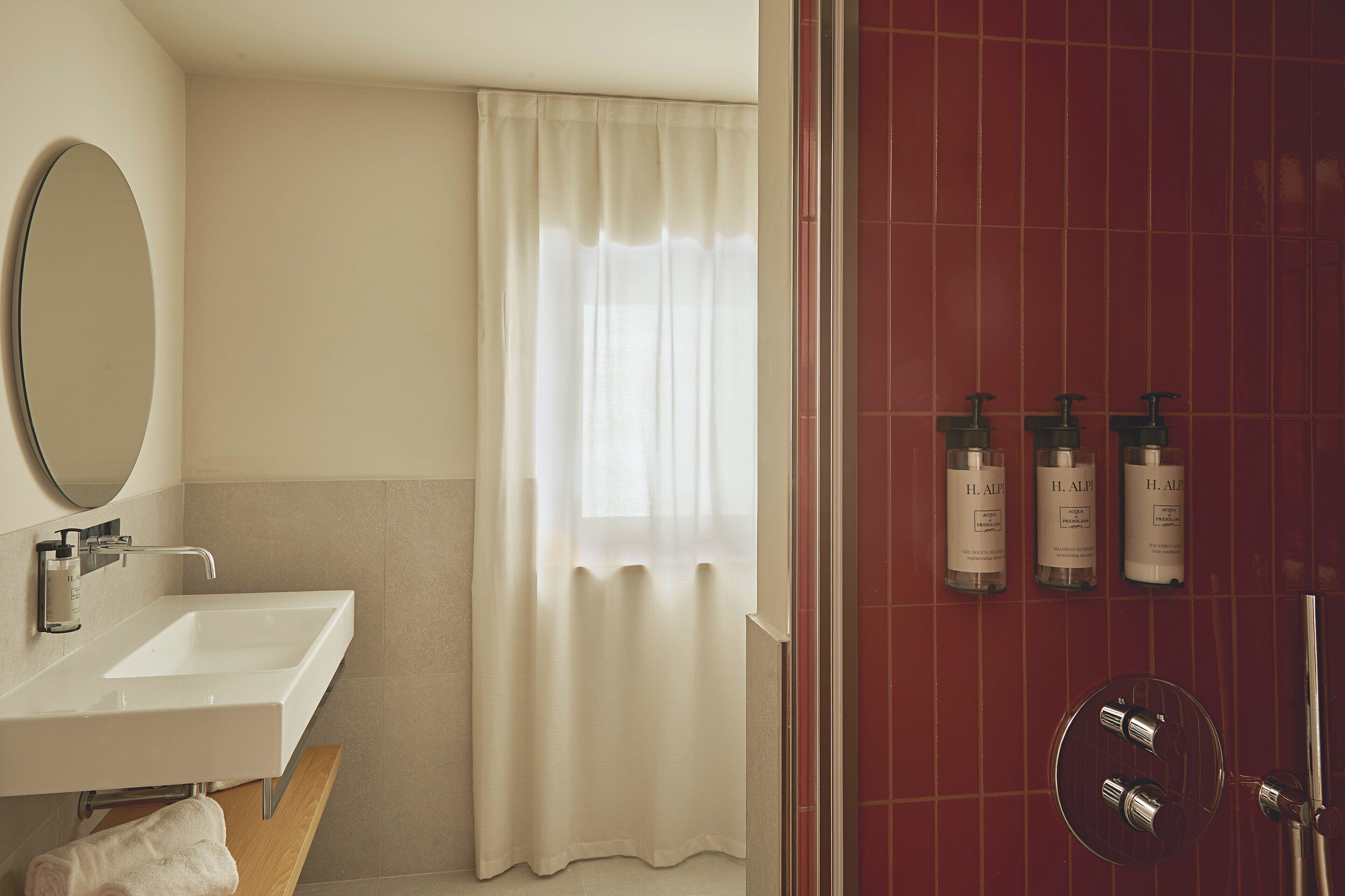 Un bagno con lavandino bianco, uno specchio, una doccia con piastrelle rosse e tre flaconi di H. ALF.