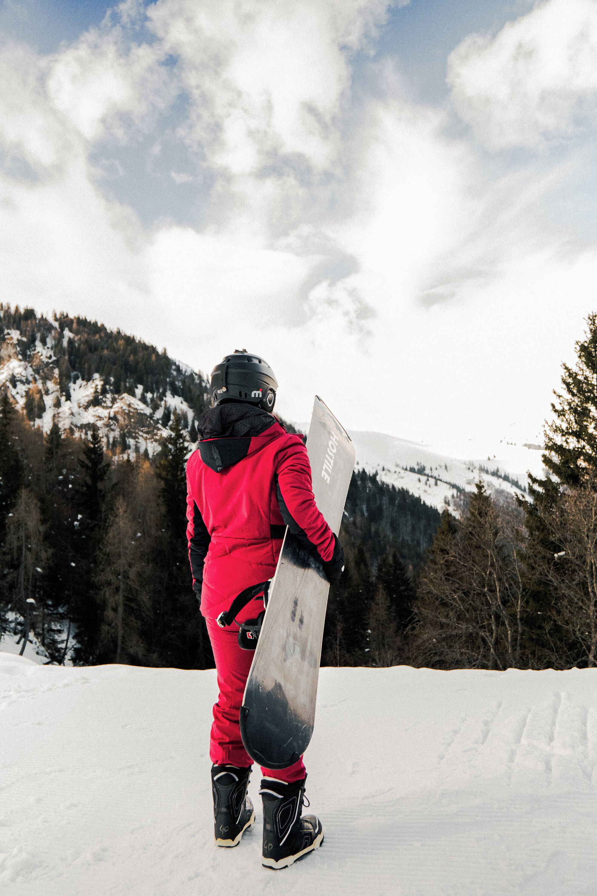 Un snowboarder con equipo rojo de pie sobre una ladera nevada con una montaña al fondo.