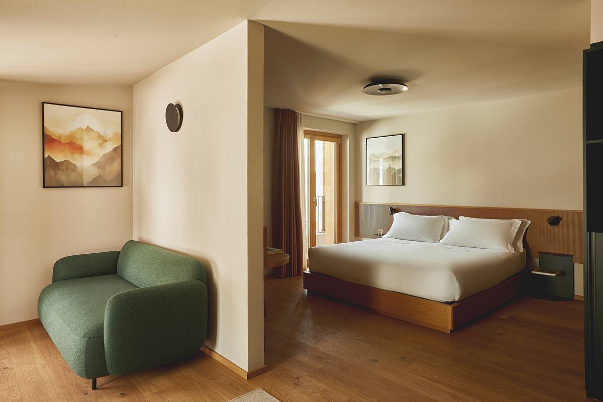 Una habitación de hotel moderna con suelo de madera, un sillón verde, una cama grande con ropa blanca y dos cuadros enmarcados en las paredes.