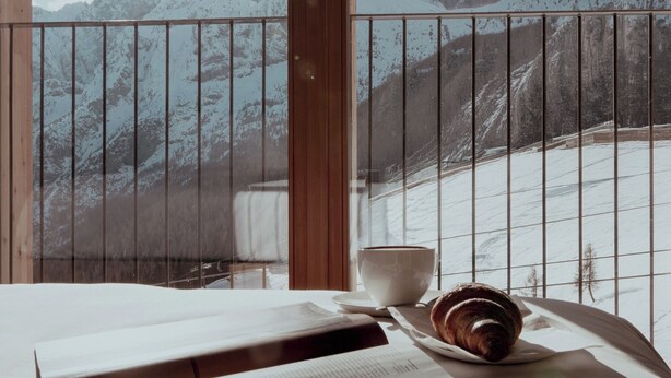 Een balkon met uitzicht op besneeuwde bergen, een tafel met een boek en croissant, en een kopje en schotel.