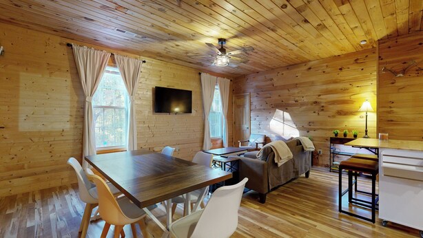 A cozy cabin interior with a wooden ceiling, a dining table with chairs, a sofa, a TV mounted on the wall, and a small kitchen area. - Hunting Home Base! 75-Acre Tennessee Getaway (Huntingdon)