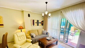 A living room with a sofa, armchair, and coffee table, a chandelier, and a view through a glass door to an outdoor area. - Dunas de Cabopino · 2BR Apartment with PRIVATE GARDEN (Marbella)