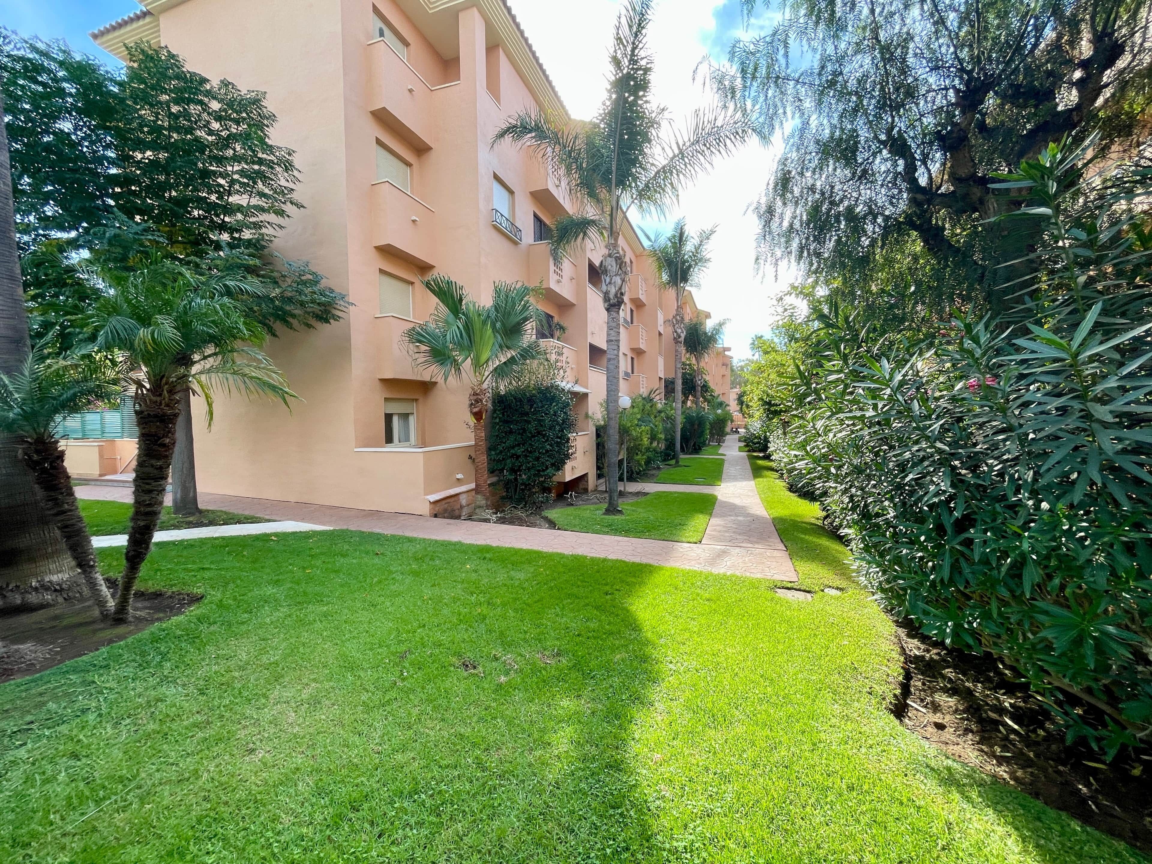Un edificio residencial con balcones, un césped cuidado y abundante vegetación circundante.