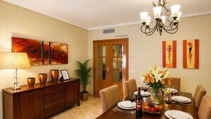 A dining room with a wooden table set for six, a chandelier, and artwork on the walls. - Dunas de Cabopino · 2BR Apartment with PRIVATE GARDEN (Marbella)