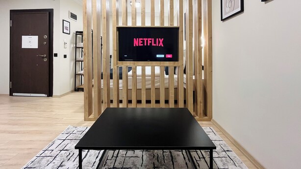 Quarto com divisória de madeira, mesa preta, TV exibindo 'NETFLIX' e quadros na parede.