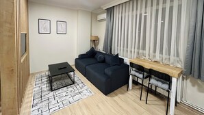 Uma sala de estar moderna com um sofá escuro, uma mesinha de centro preta, uma escrivaninha de madeira com duas cadeiras e uma grande janela com cortinas leves.
