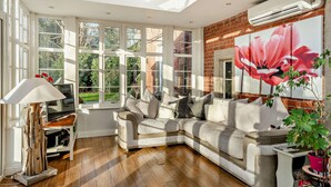 A sunlit room with a sectional sofa, a TV on a wooden stand, and a large window with a view of greenery. - Luxury Detached House,  Private Grounds. With Hot Tub, Sauna, Pool Table. (Weston)