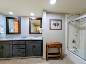 A bathroom with a double vanity, two mirrors, a framed animal picture, a small wooden side table, and a walk-in shower. - Lake Views, Sleeps 6, New Listing! (Keystone)