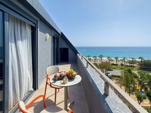 Un balcó amb taula preparada per a dos, amb vista a una platja de palmeres i un cel blau clar.