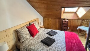 Un dormitorio con techo inclinado, paredes de madera, cama con ropa de cama estampada, almohada roja y mesita de noche con lámpara.