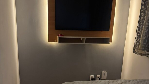 A modern hotel room with a large flat-screen TV mounted on the wall, a bed with a grey quilt, and a wooden floor. - Studio Paris - Downtown Niterói (Niterói)