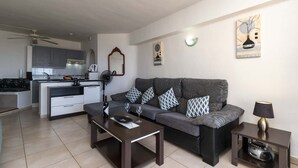 Una sala de estar moderna con un sofá gris, una mesa de centro y una mesita auxiliar con una lámpara. Se ve una cocina al fondo con gabinetes grises y una encimera blanca.