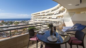 Un balcón con mesa lista para comer, con vista a una zona costera con varios edificios y un cielo azul despejado.
