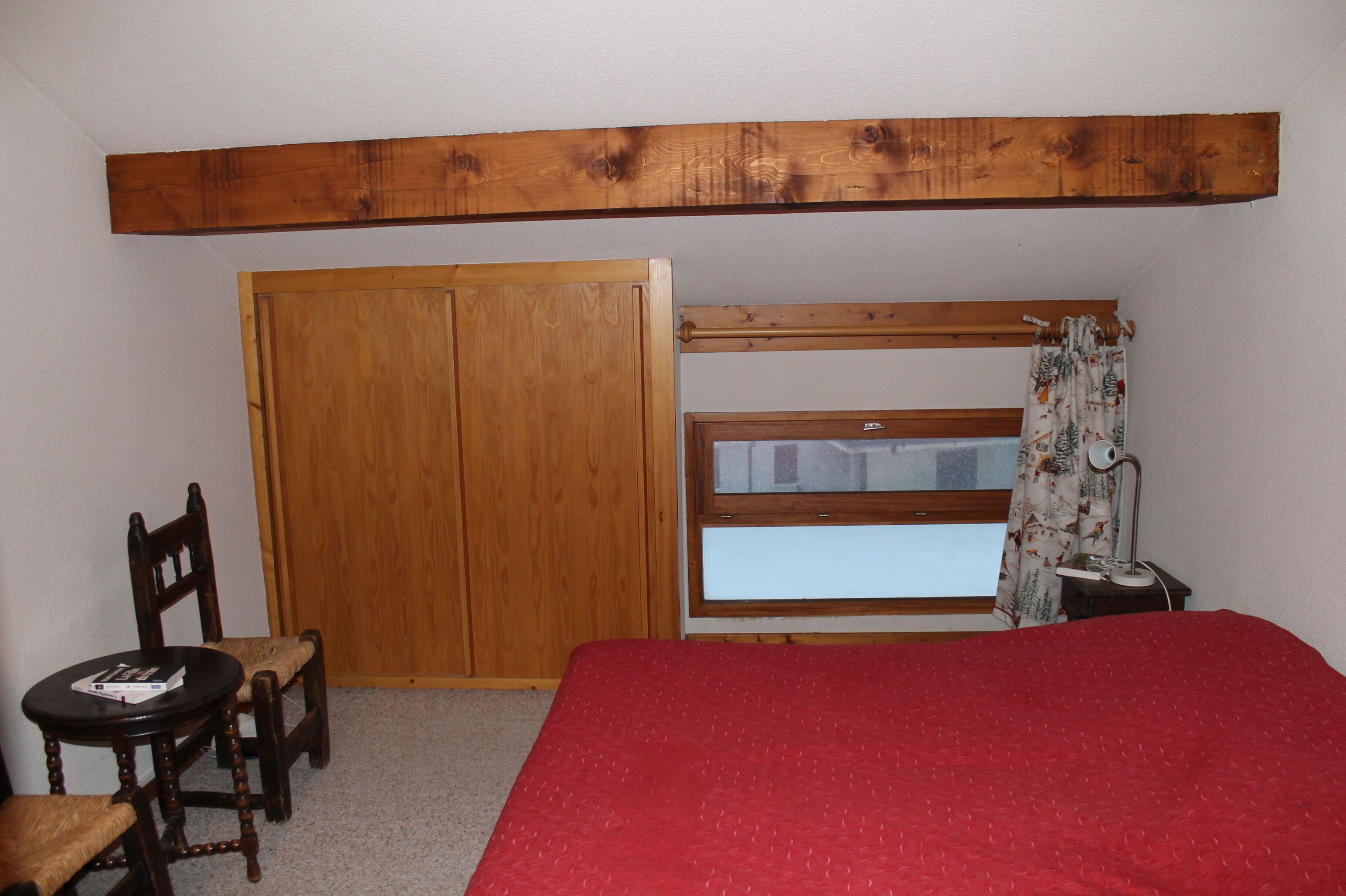 A bedroom with a red bedspread, wooden wardrobe, and a window with curtains.