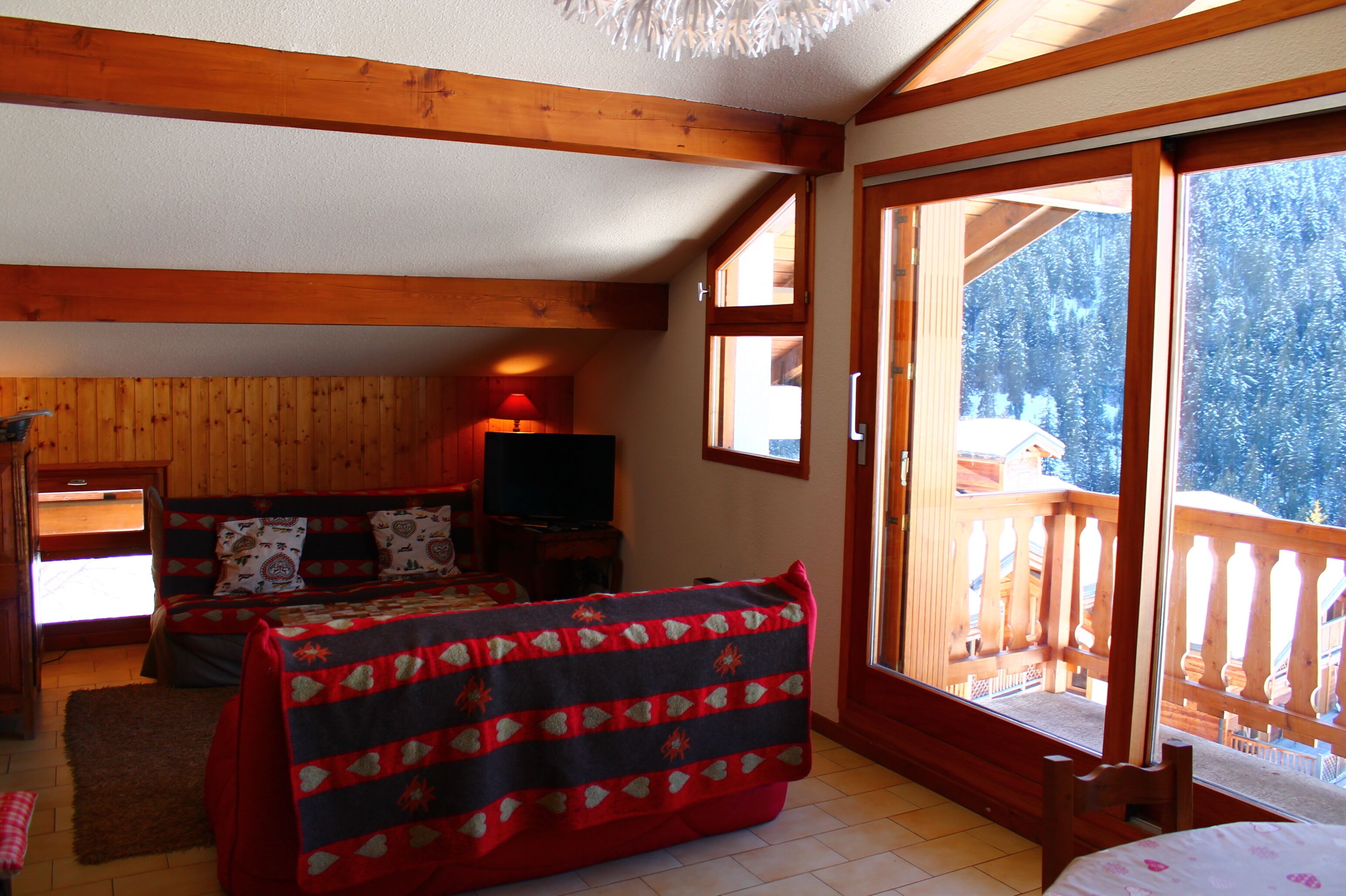A cozy room with wooden beams, a bed with a patterned blanket, a TV, and a balcony with a wooden railing.