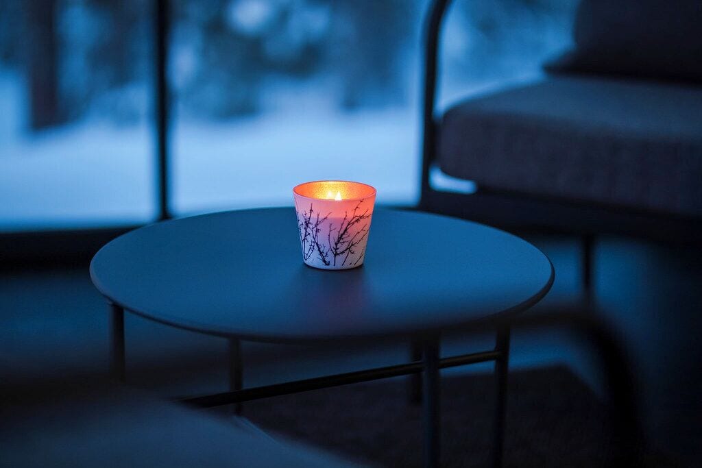 A candle on a round table in a dimly lit room with a view of a snowy landscape through a window.