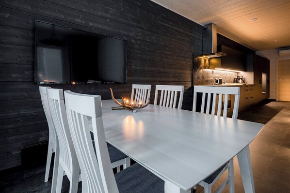 Un espace repas moderne avec une table et des chaises blanches, un téléviseur à écran plat fixé au mur, et un coin cuisine doté d’un plan de travail et d’appareils électroménagers.