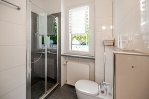 A bathroom with a glass shower enclosure, a white toilet, a window with a white curtain, and a towel rack. - ApartmentsGarda - Albisano Sky House (Torri del Benaco)