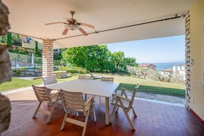 An outdoor patio with a table and chairs, a ceiling fan, and a view of a green landscape and buildings. - ApartmentsGarda - Albisano Sky House (Torri del Benaco)