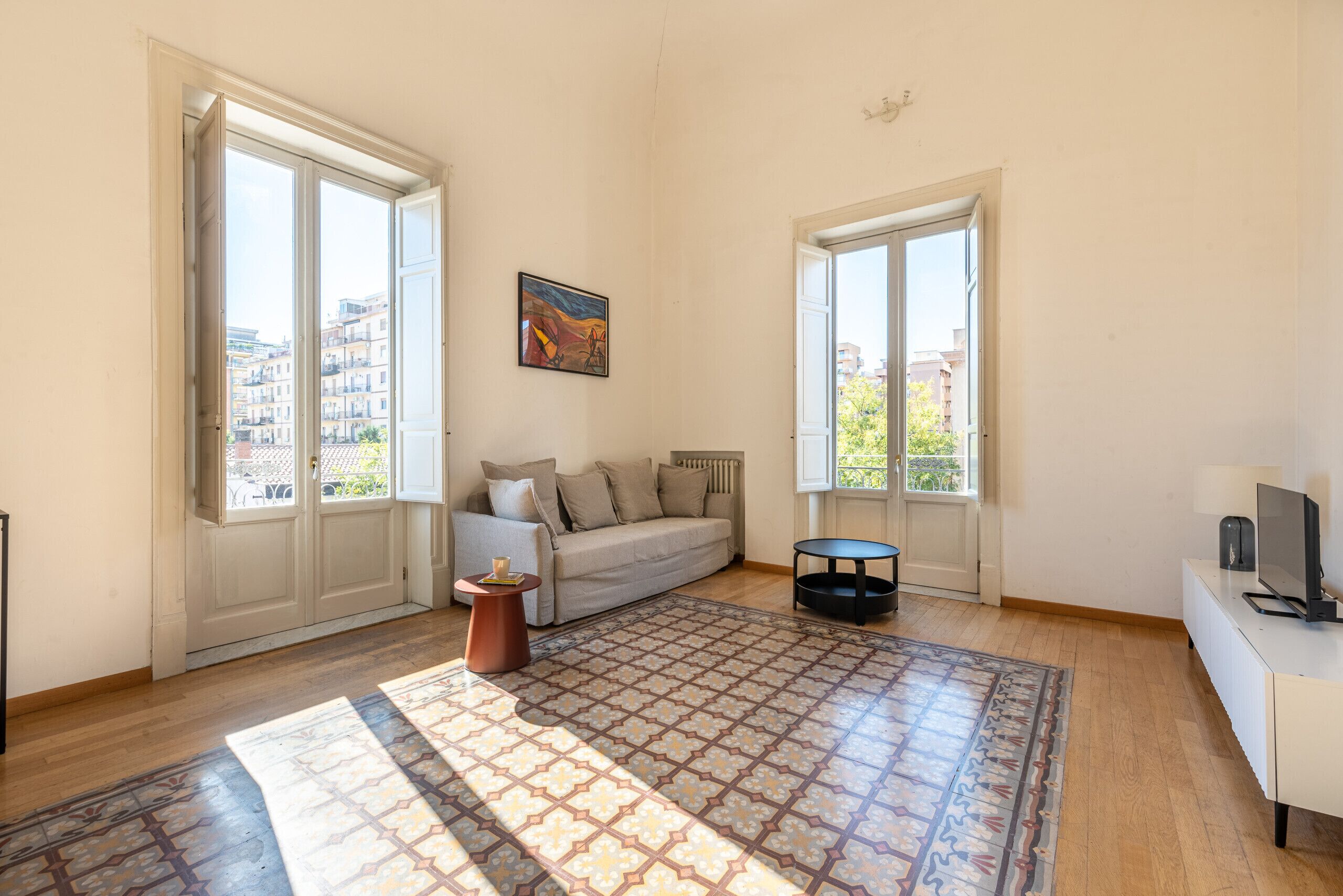 A spacious living room with a sofa, a coffee table, and a patterned rug. There are two windows with views of buildings outside.