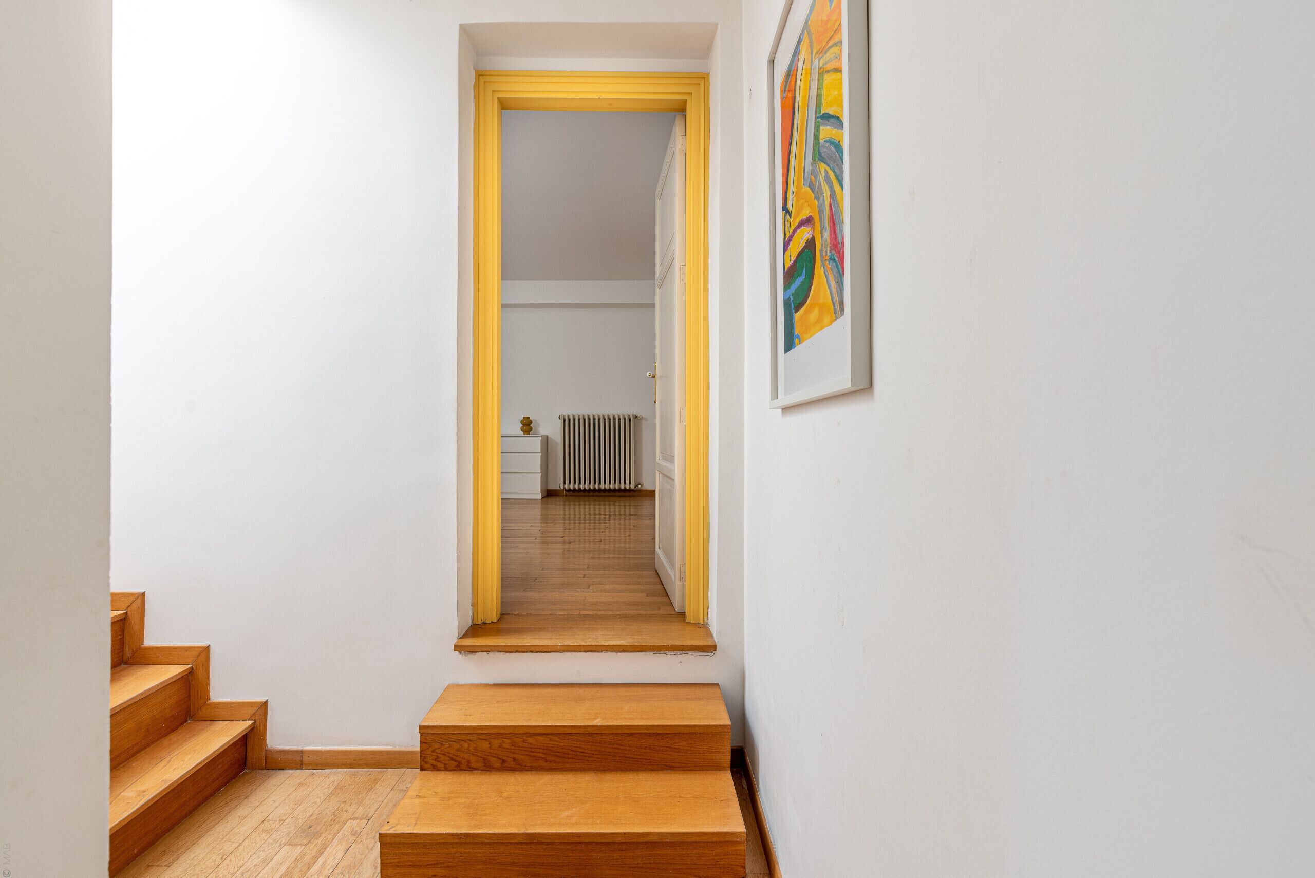 A narrow hallway with wooden steps leading to a room with a radiator and a framed artwork on the wall.
