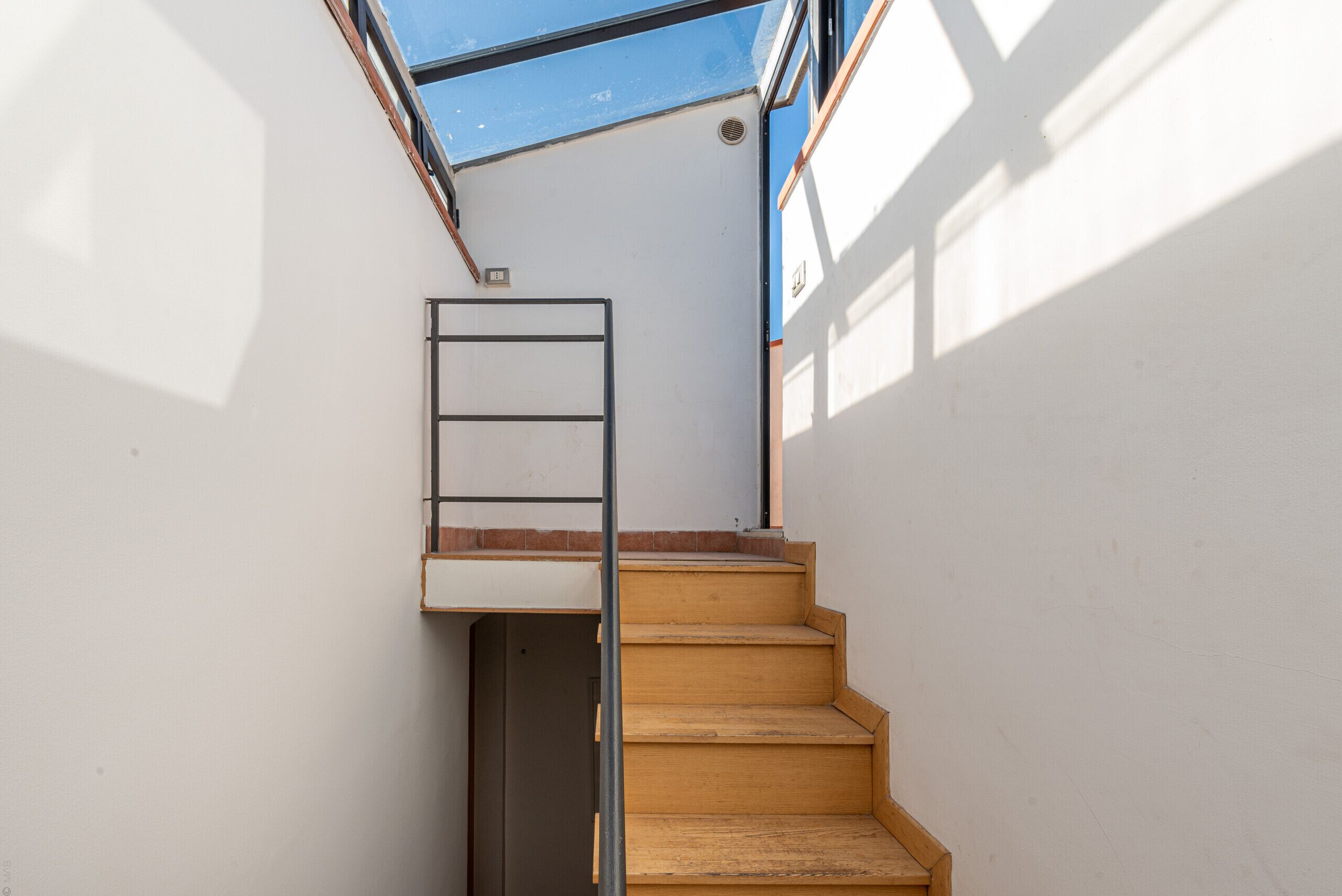 A narrow staircase with wooden steps and a metal handrail, leading to a landing with a small window.