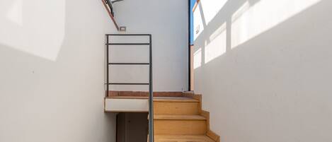 A narrow staircase with wooden steps and a metal handrail, leading to a landing with a small window.