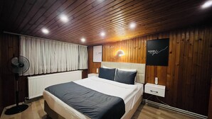 Quarto com teto de madeira, cama com roupa de cama branca e escura, um ventilador, um radiador e um aplique de parede.