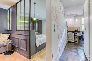 Ein modernes Schlafzimmer mit Bett, Sofa und einem kleinen Tisch. Es gibt eine Küchenzeile mit Barhocker und einem Kühlschrank.