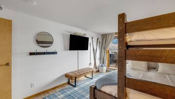 A bedroom with a bunk bed, a flat-screen TV mounted on the wall, a bench, and a window with curtains.