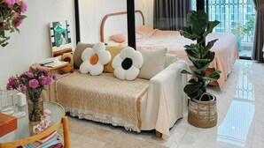A modern bedroom with a large bed, a bedside table with a vase of flowers, a small table with books, and a large window with curtains.