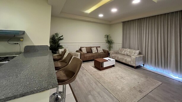 Una sala de estar moderna con área de cocina, que incluye un taburete, un conjunto de sofás y un amplio ventanal con cortinas.