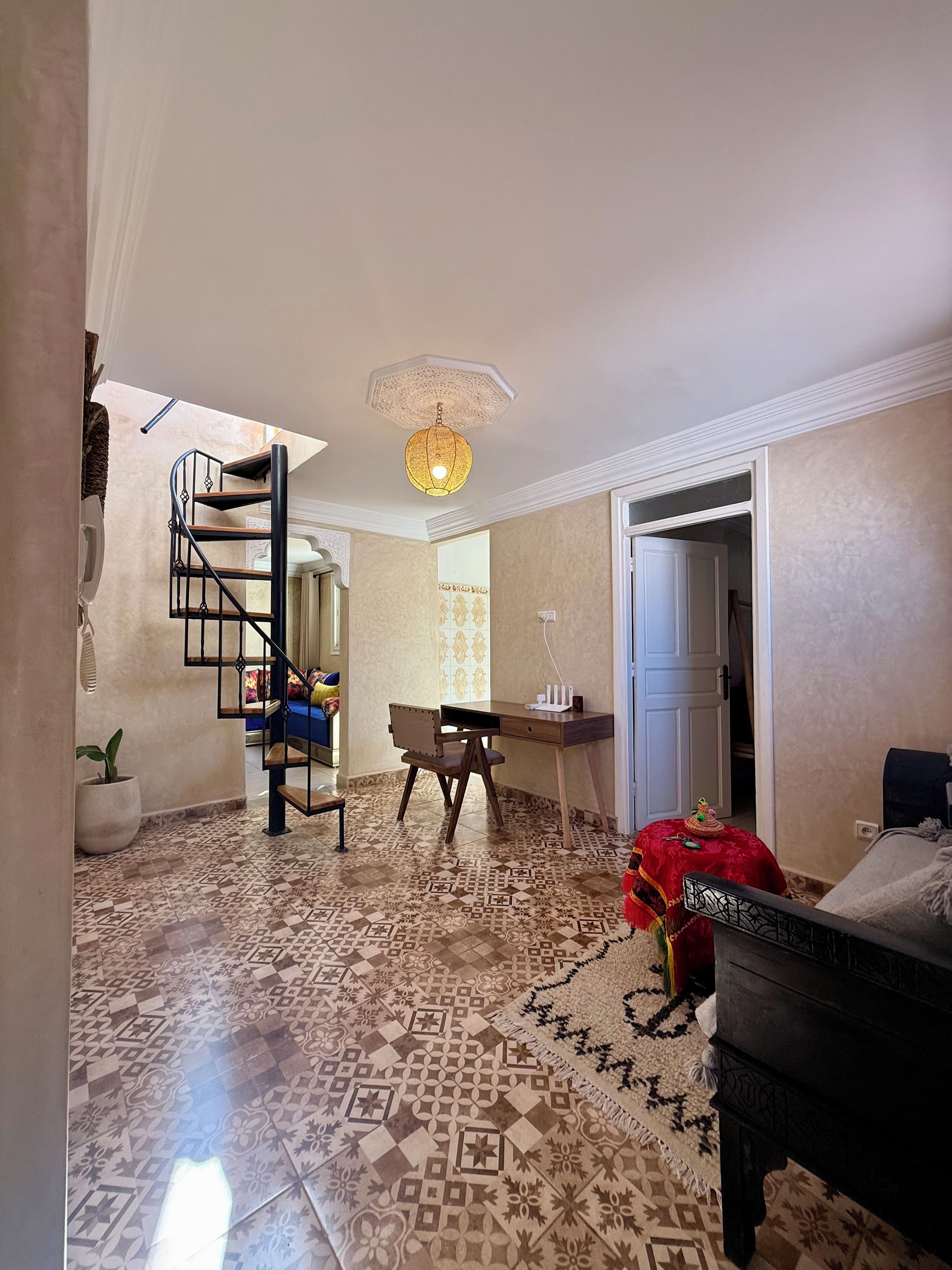 Une pièce avec un sol à motifs, un bureau, une chaise, un lit, un escalier et un luminaire suspendu.