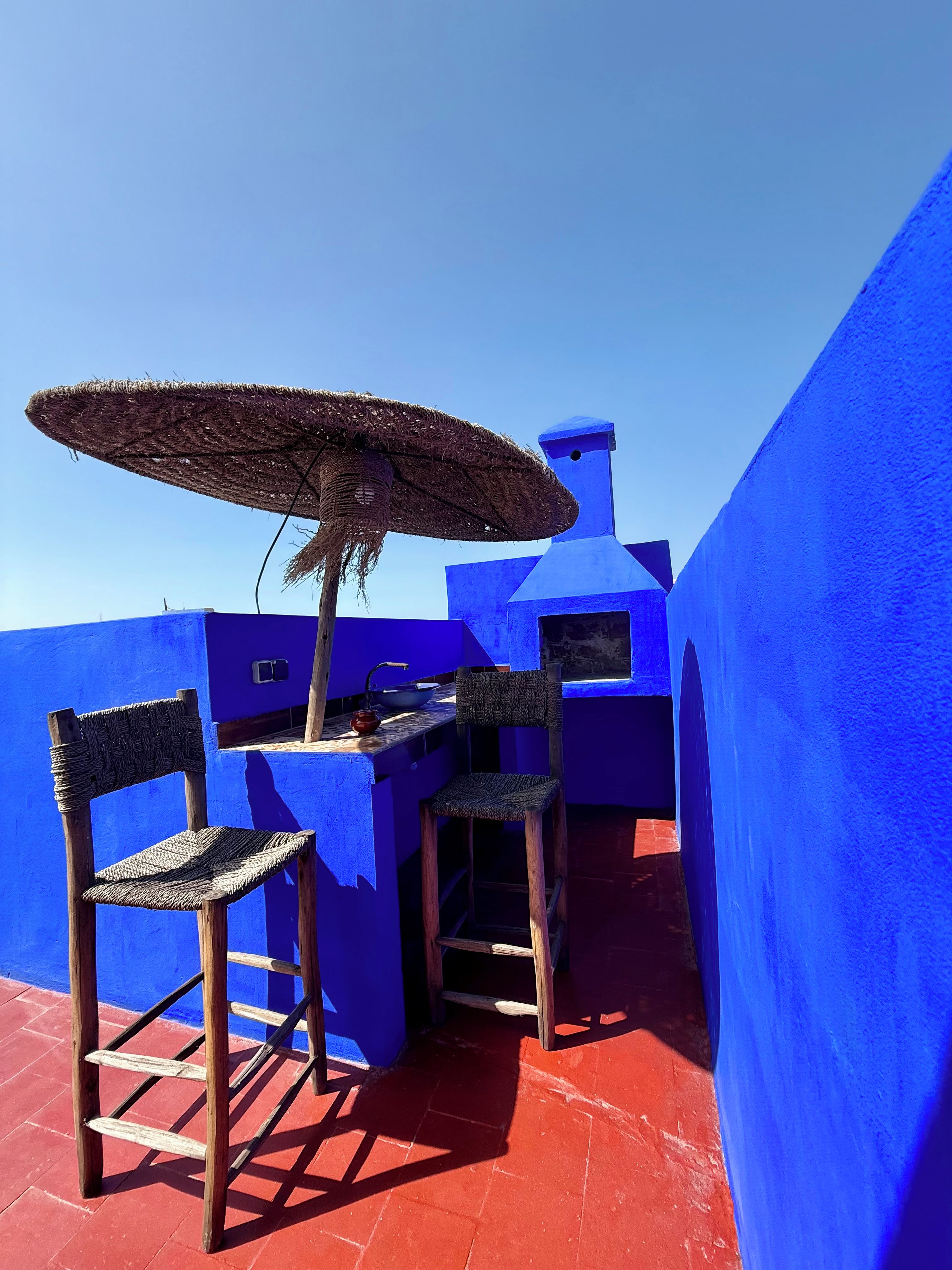 A blue-painted outdoor seating area with a woven umbrella and wooden chairs.