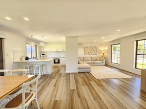 Una cocina moderna con armarios blancos, un comedor con mesa de madera y sillas blancas, un sofá y un amplio ventanal.