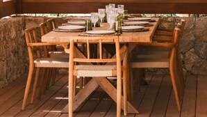 Un tavolo da pranzo in legno apparecchiato per quattro, illuminato da lampade a sospensione intrecciate, circondato da pareti in pietra e ampie finestre con persiane in legno.