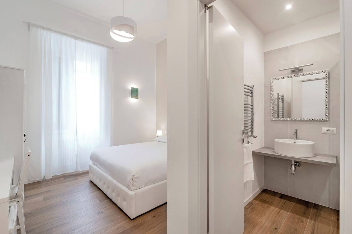 A bedroom with a bed, a bedside table, and a window with curtains. The room has a wooden floor and white walls.