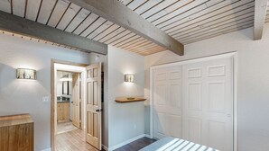 Un dormitorio con techo de madera, una cama con ropa de cama blanca, una mesita de noche, una estantería con un cuenco y una puerta que da al baño.