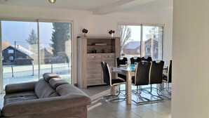 A modern living room with a sofa, dining table, and chairs. There is a large window with a view of a snowy landscape.