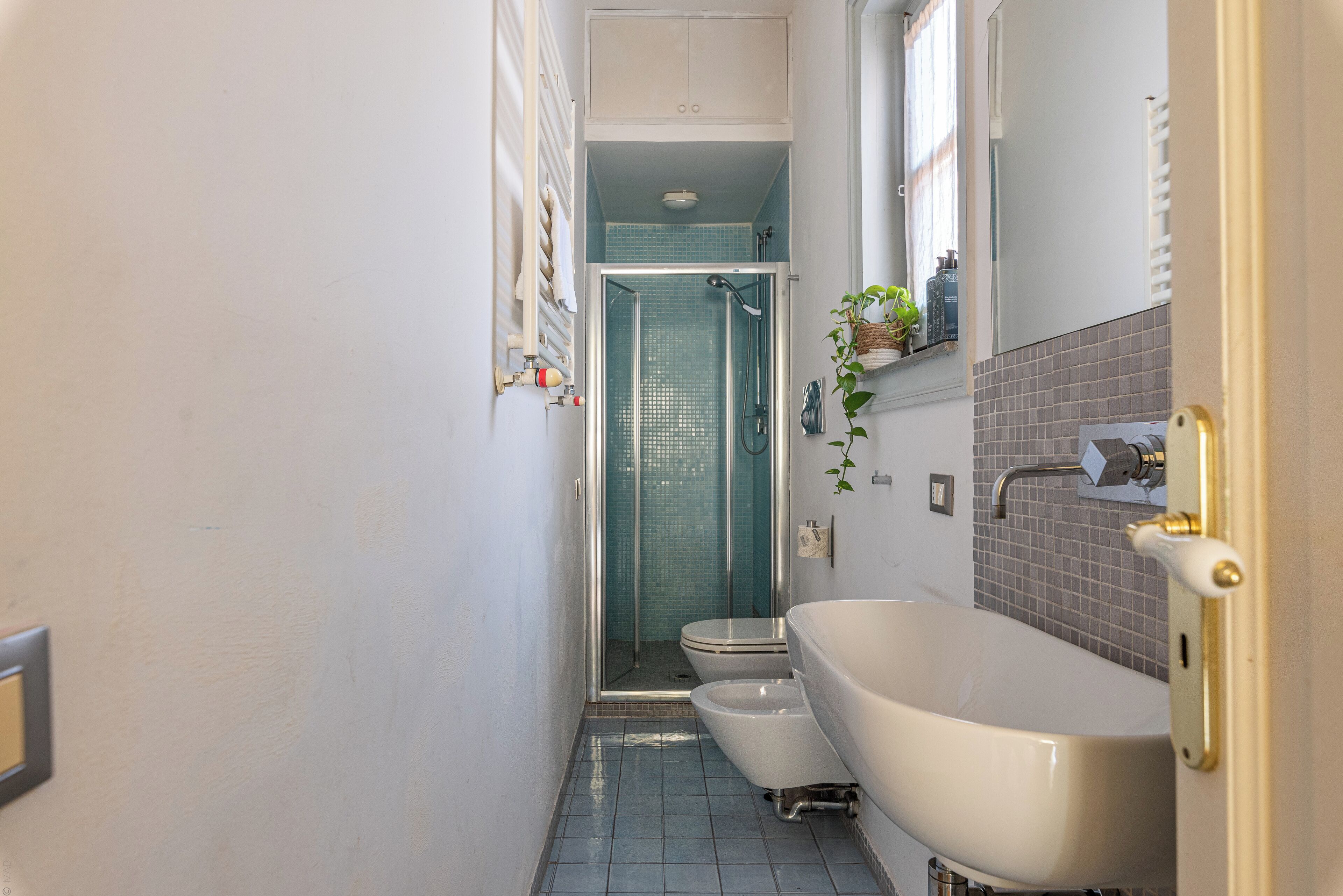 Un baño estrecho con una bañera blanca, dos inodoros blancos y una ducha con mampara de vidrio.