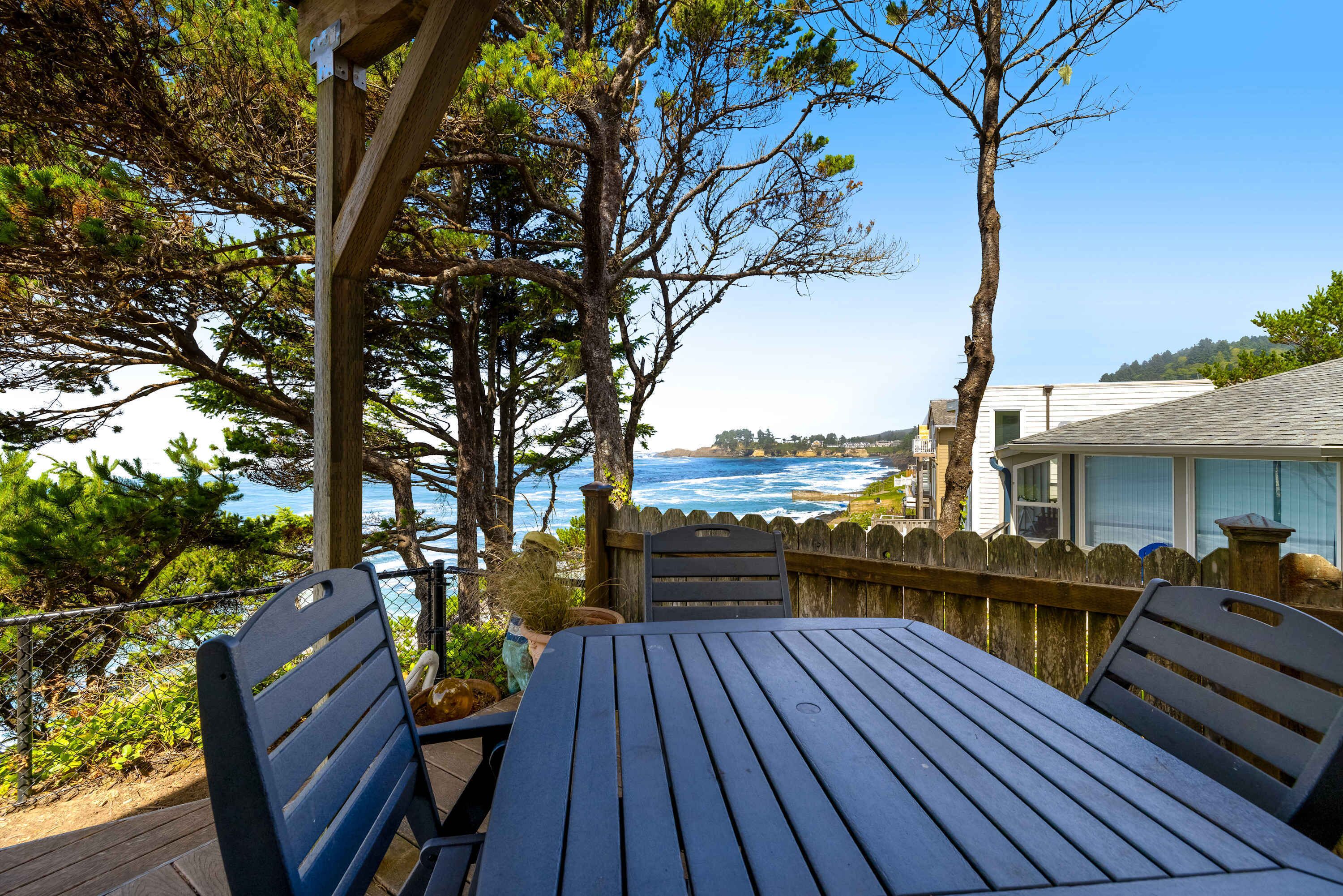 A wooden deck with a table and chairs, overlooking a coastal area with trees and a view of the ocean.