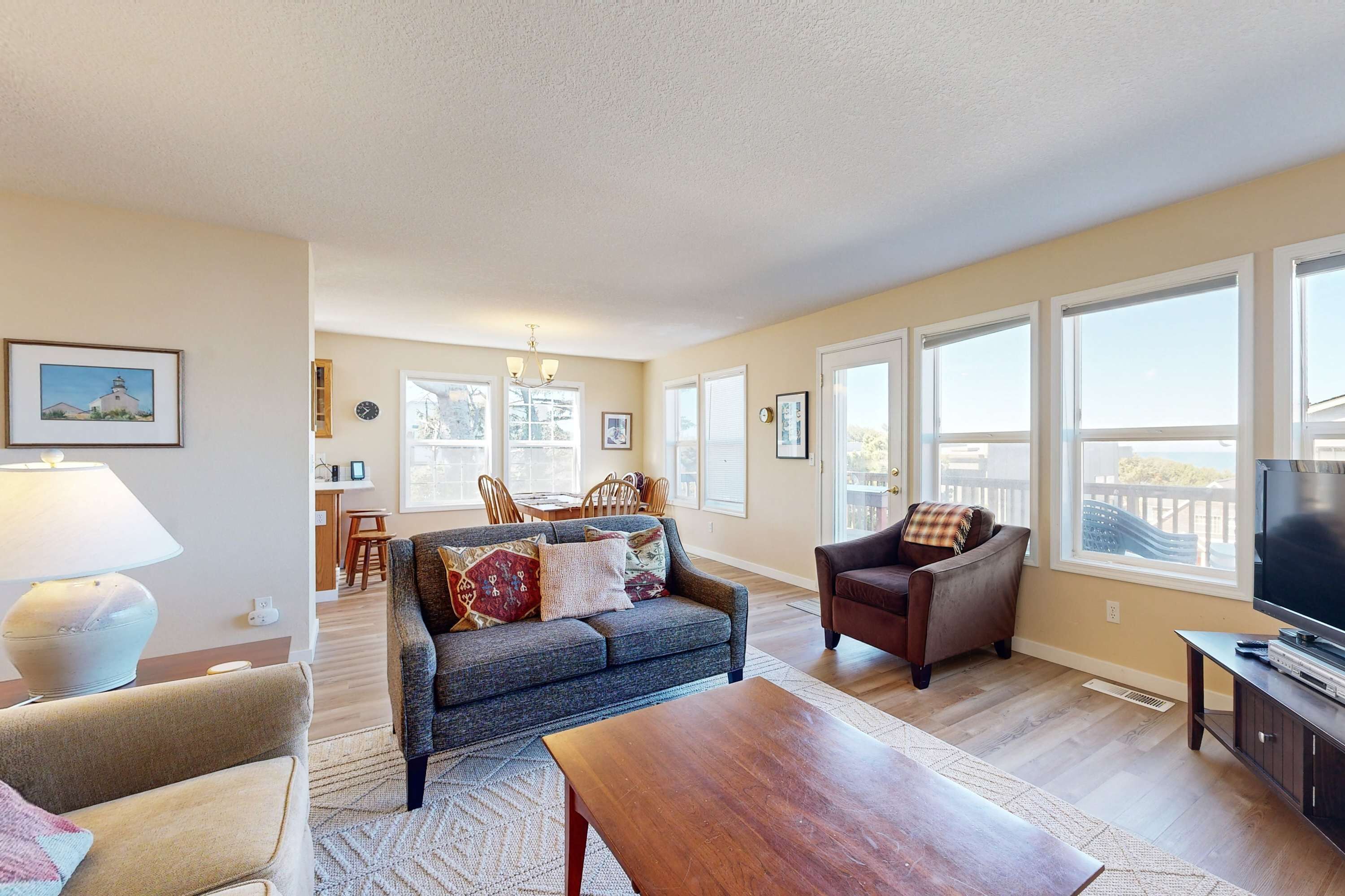 A spacious living room with a sofa, armchair, and a wooden coffee table. There are large windows offering a view of the outdoors.